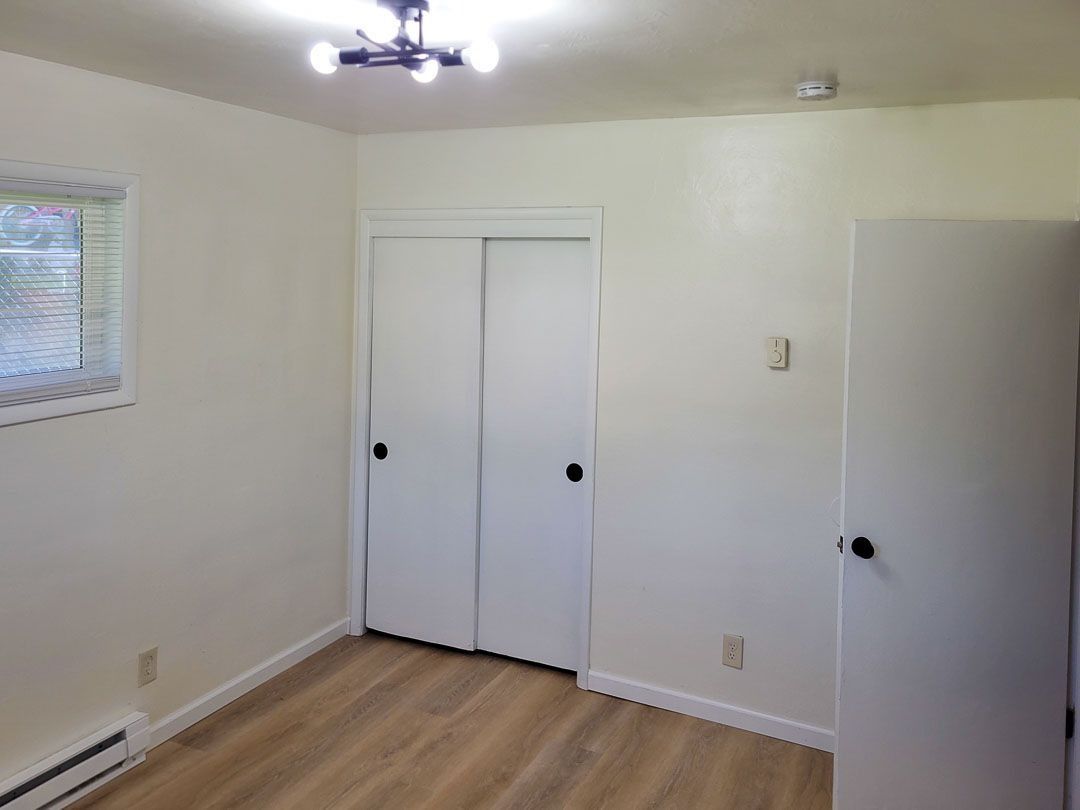 Empty bedroom with white closet doors, wood floor, and a small window.