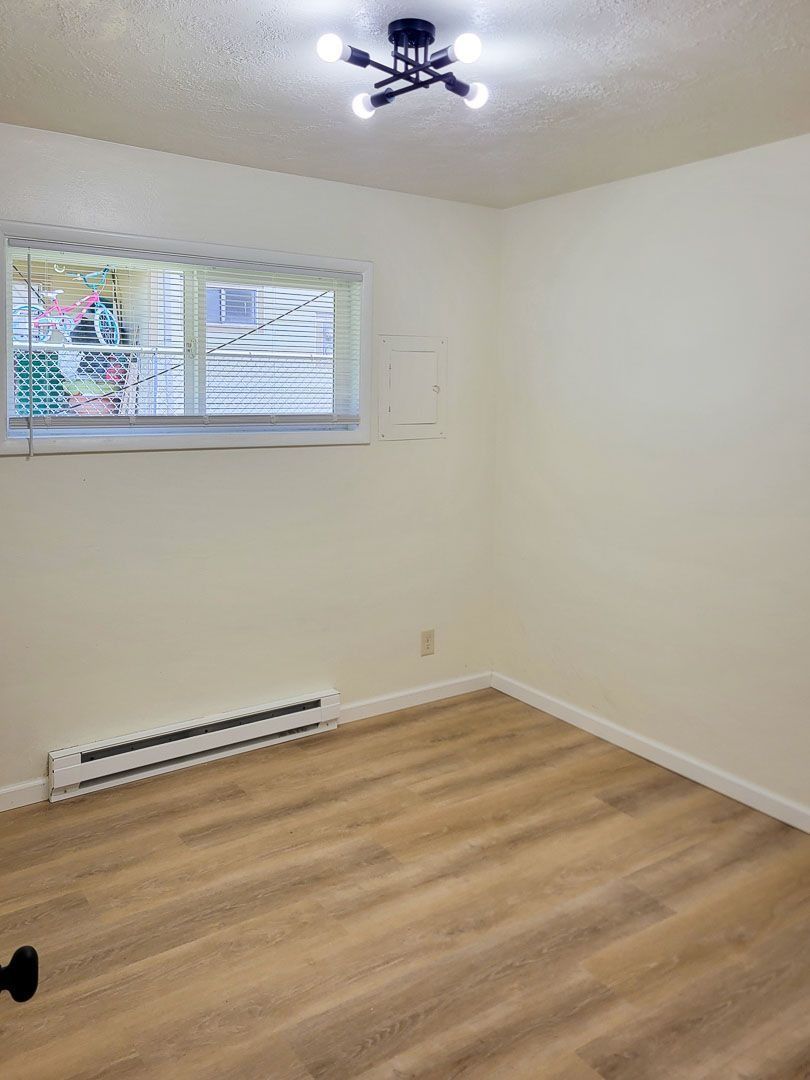 Empty room with white walls, wood flooring, a window, ceiling light, and baseboard heater