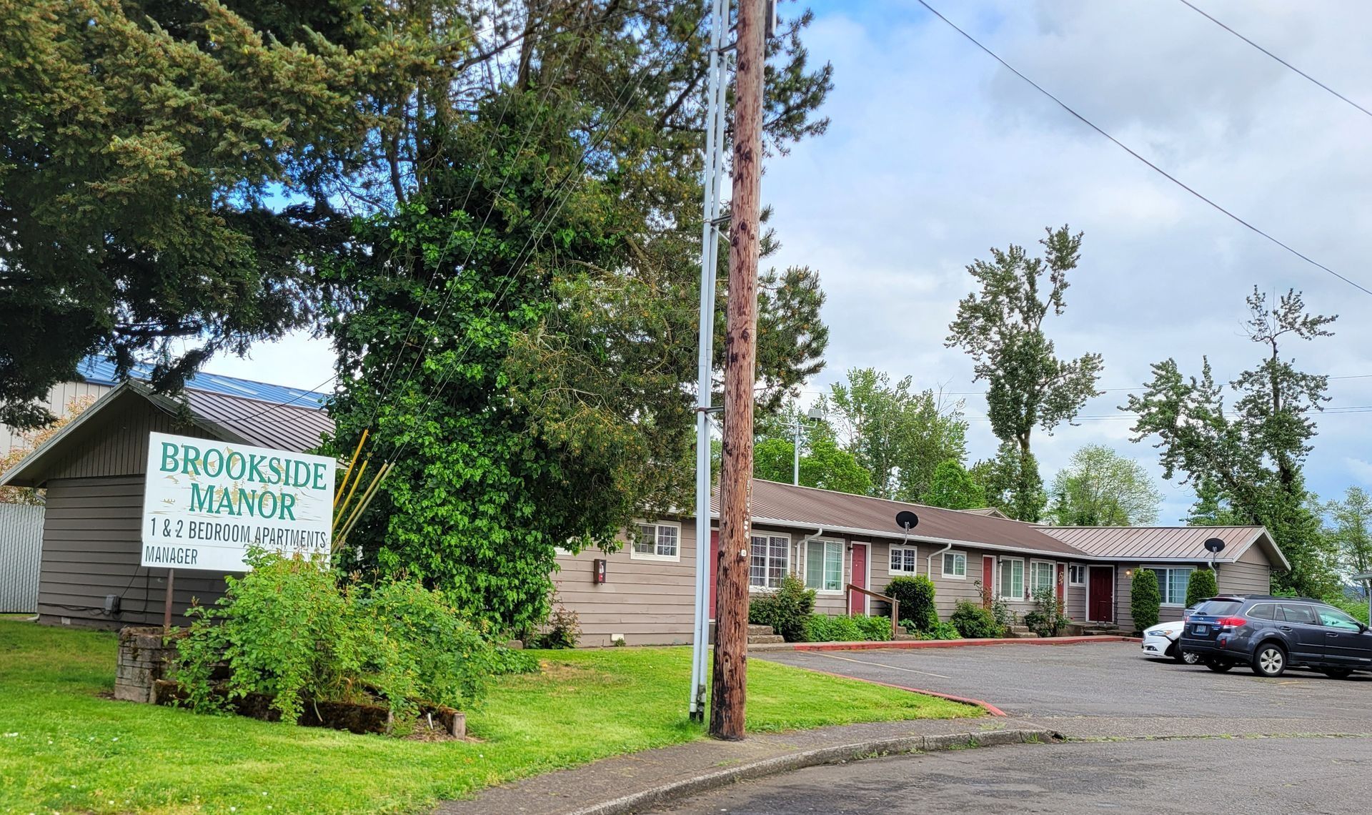 Brookside Apartments sign beside a tan apartment building, with trees and parked cars on a cloudy day
