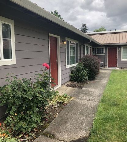 Apartment walkway beside gray buildings with red doors and small shrubs under a cloudy sky