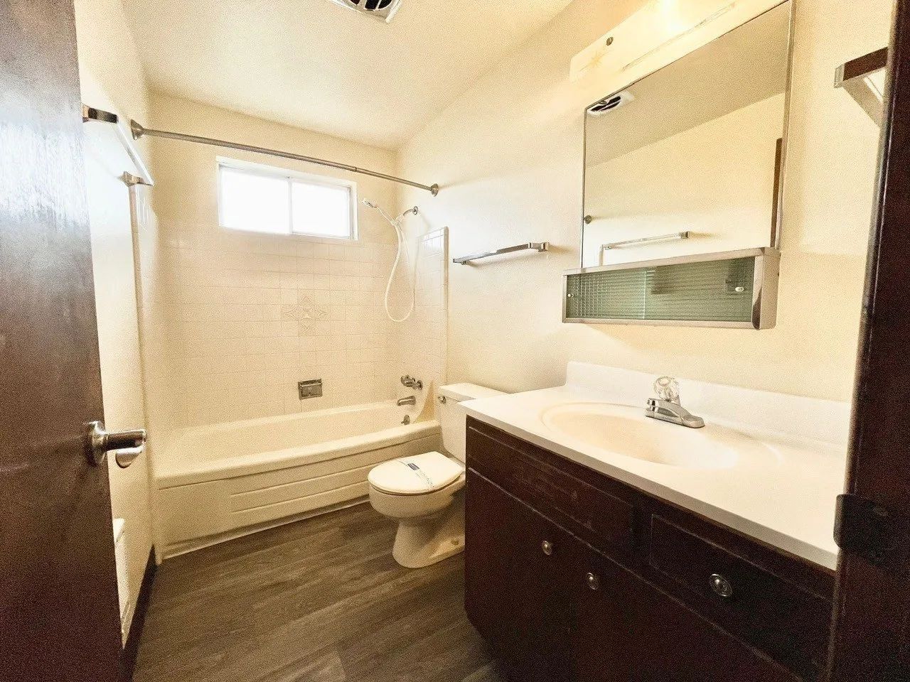 Small beige bathroom with bathtub, toilet, dark vanity, mirror, and window.