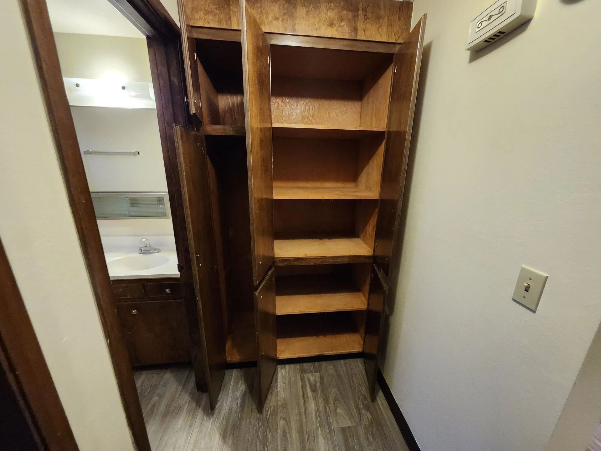 Small wooden closet with shelves and hanging mirror beside a bathroom doorway