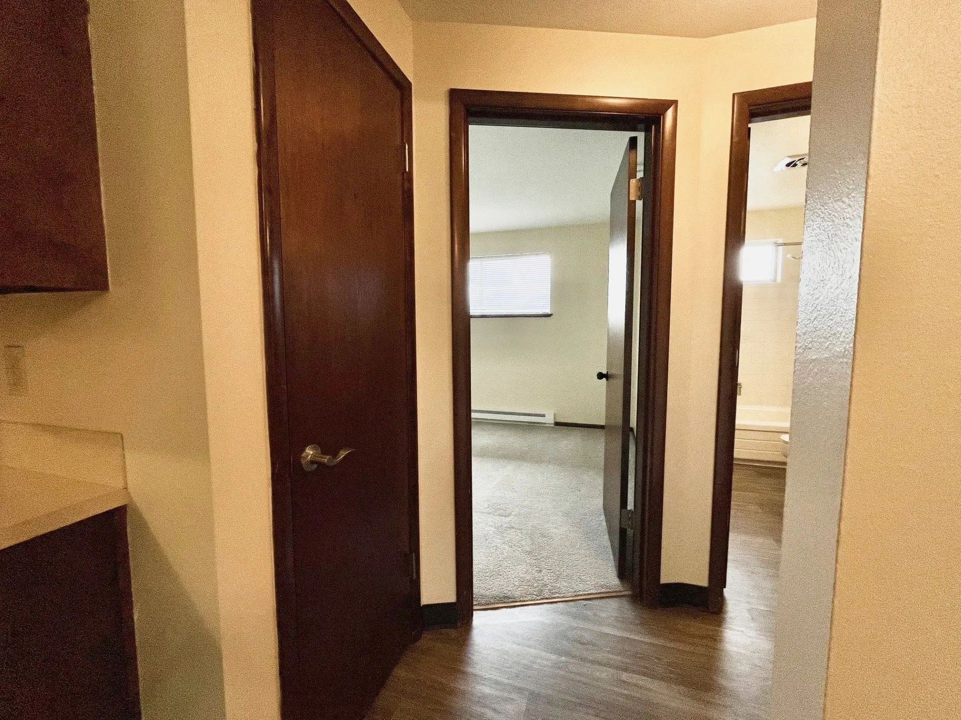 Hallway with wood floors, beige walls, and open doorways leading to a carpeted room.
