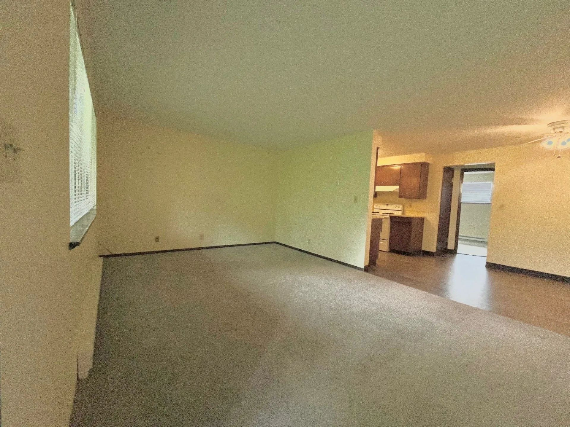 Empty carpeted living room with beige walls and open view into a kitchen area.