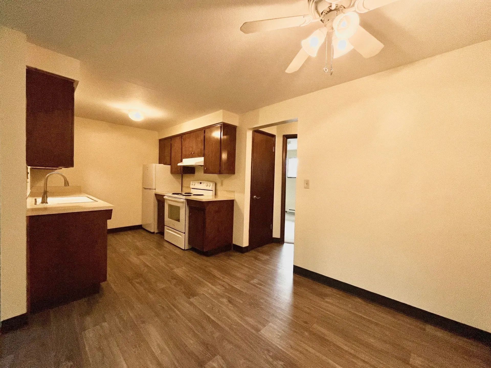 Empty kitchen-living area with wood floors, white walls, dark cabinets, and ceiling fan.