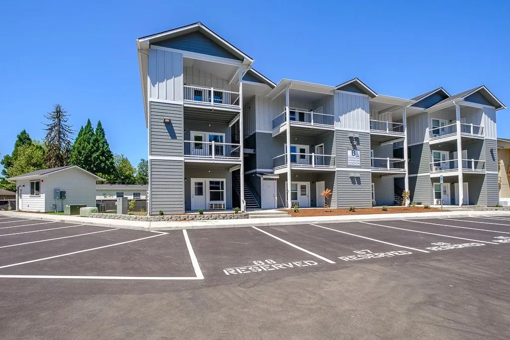 Modern three-story apartment building with balconies and parking lot under a clear blue sky