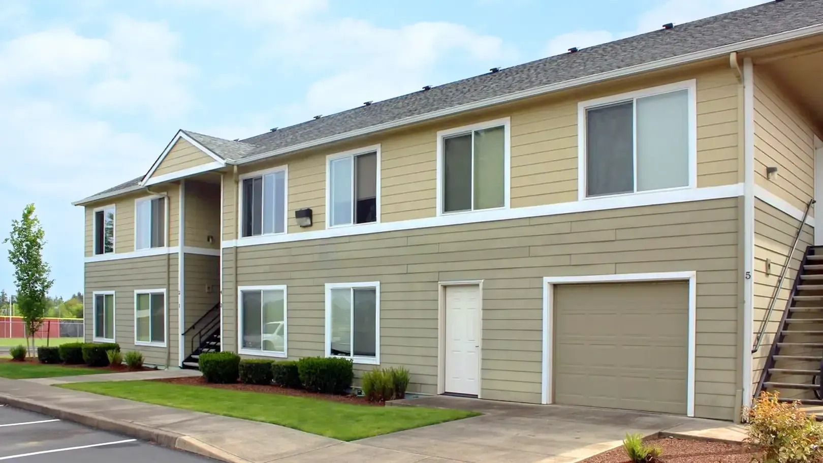 Two-story beige apartment building with white trim, balconies, garage, and parking lot under cloudy sky
