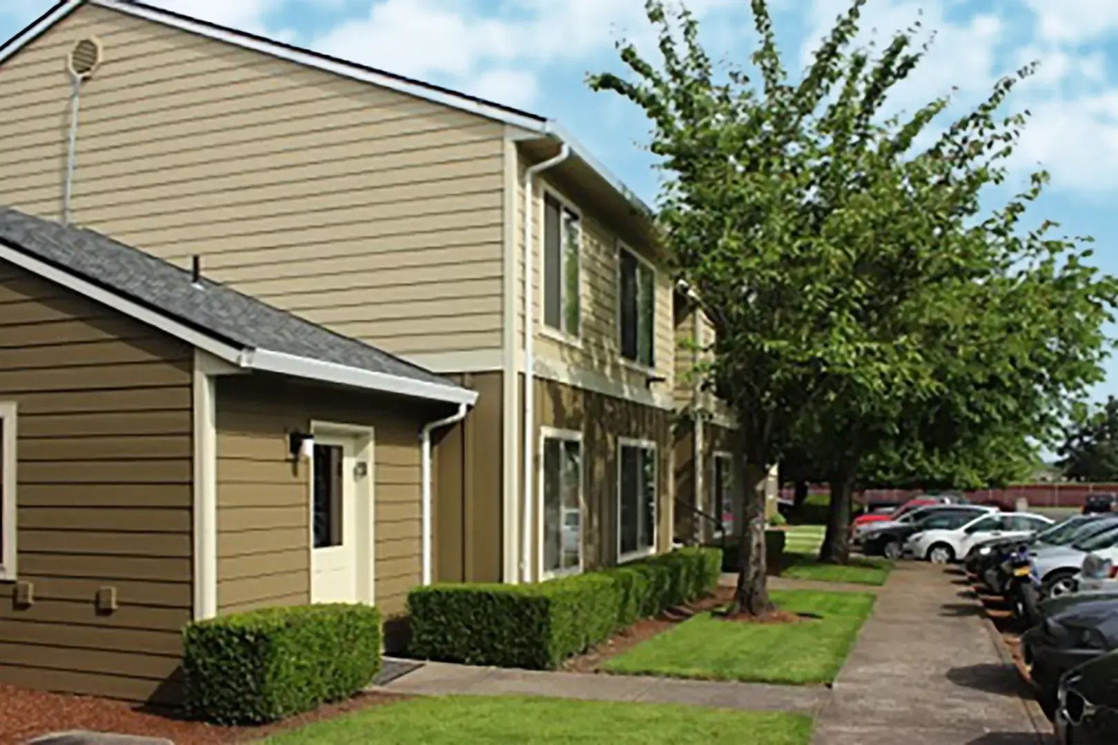 Apartment complex with beige buildings, hedges, sidewalk, and parked cars along the street