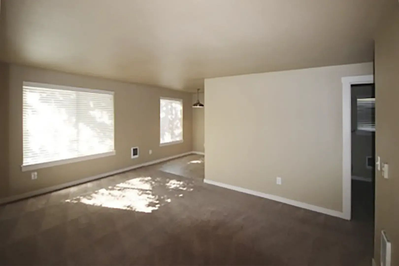 Empty beige carpeted living room with white walls, large window, and sunlight on the floor