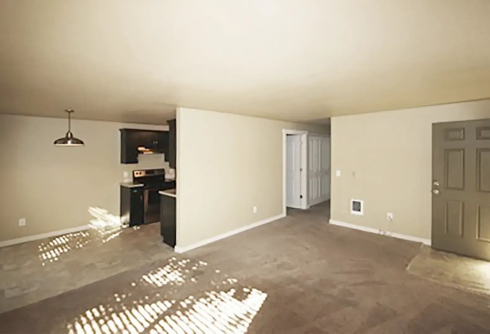 Empty carpeted living room with beige walls, open doorway, and sunlit floor near a kitchen nook