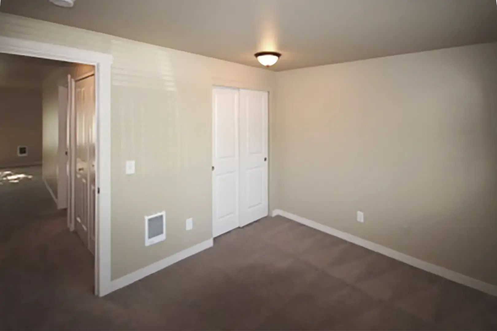 Empty beige bedroom with carpet, white closet doors, and hallway visible through an open doorway