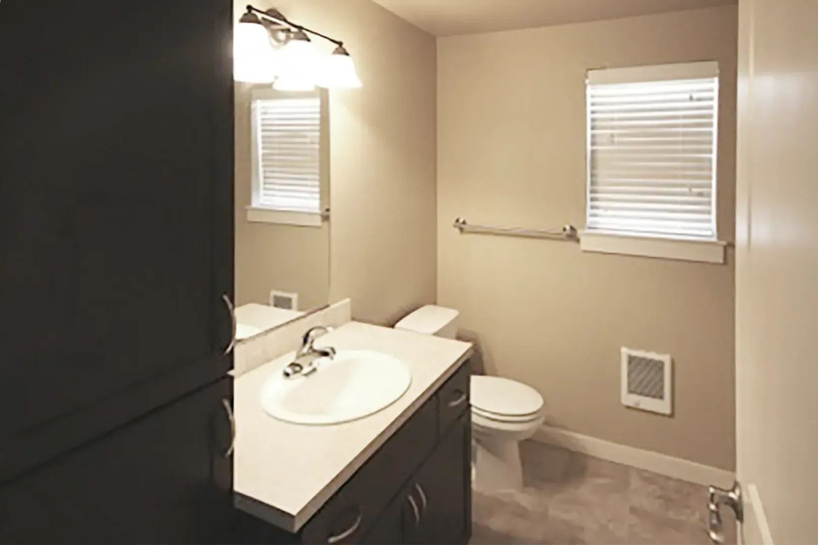 Compact beige bathroom with white sink, toilet, wall towel bar, and two small windows.