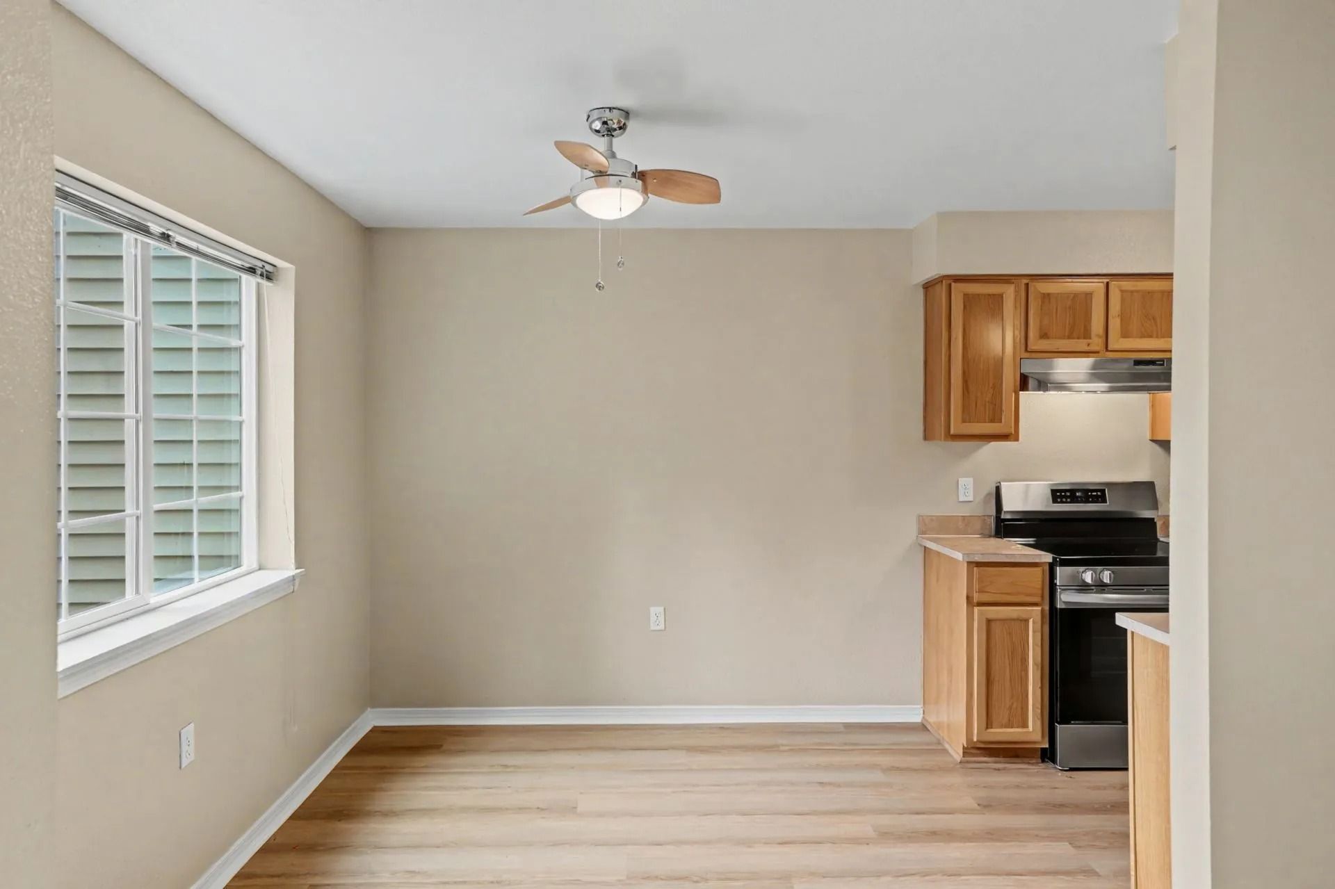 Empty beige room with wood floor, window blinds, ceiling fan, and a small kitchen area on the right