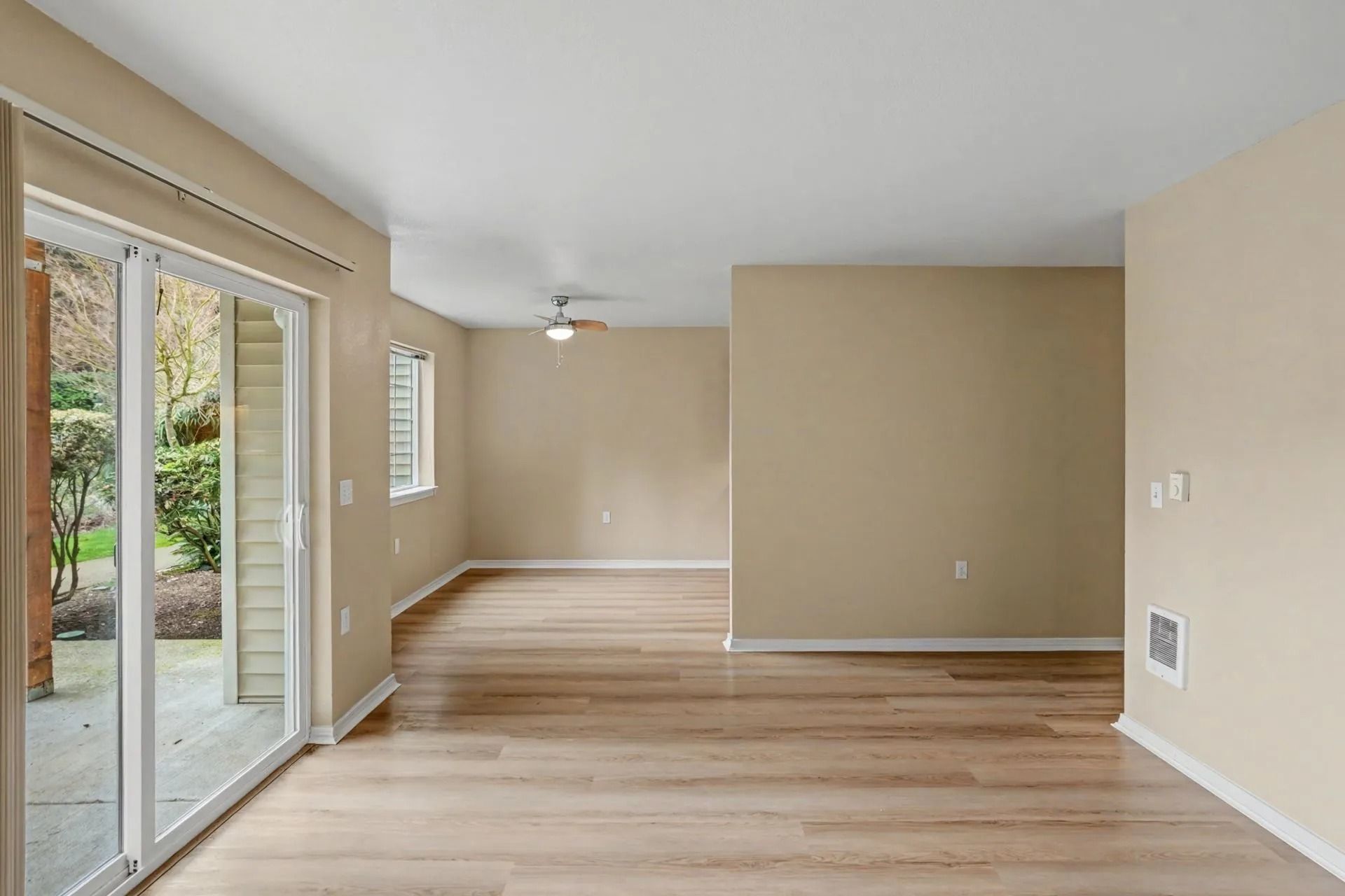 Empty beige living room with sliding glass door, light wood floors, and ceiling fan