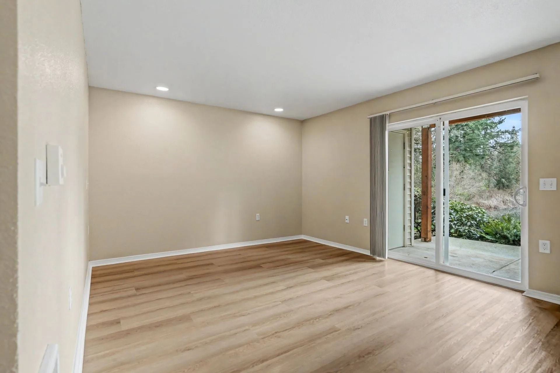 Empty beige room with light wood floors and sliding glass doors opening to a green yard