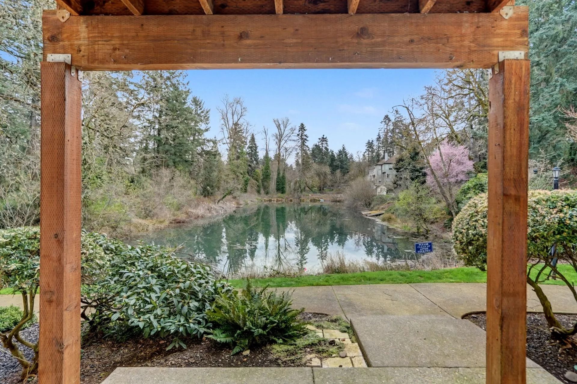 Wooden frame overlooks a calm pond, trees, and a garden path under a clear blue sky.