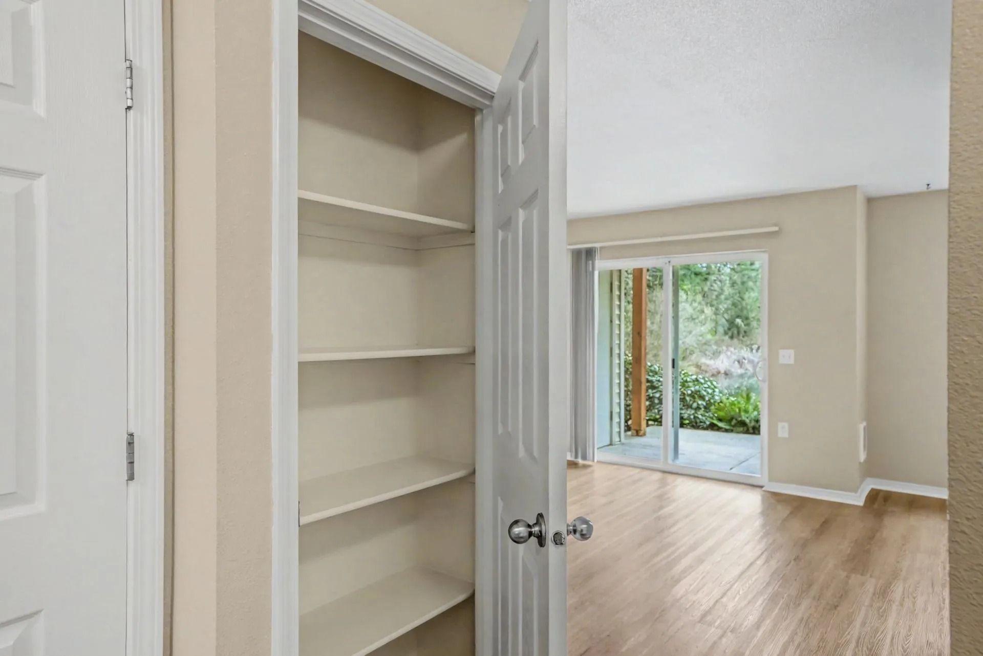 Open closet with shelves beside a bright empty room and sliding glass door to greenery