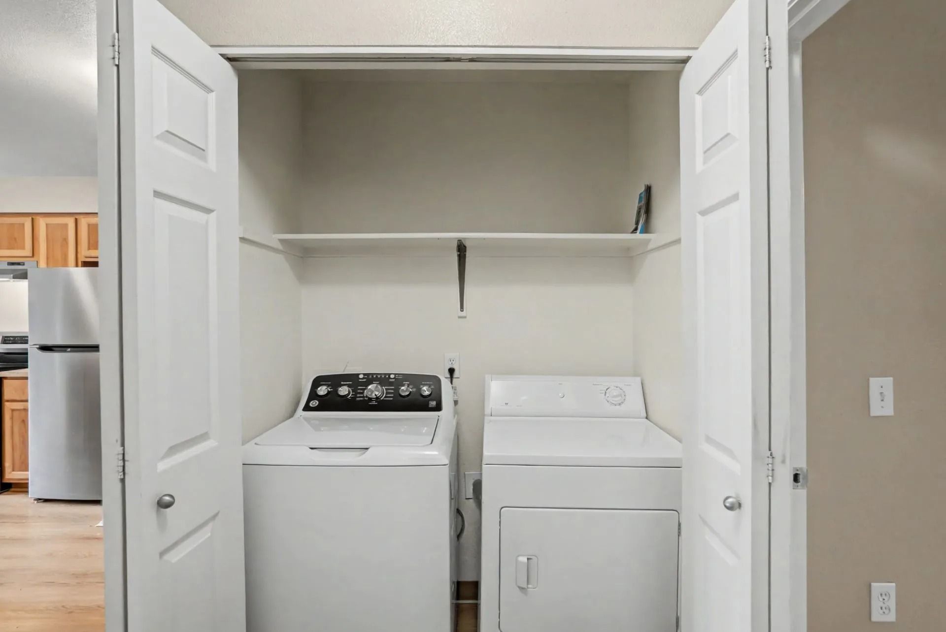 Laundry closet with a washer and dryer under a shelf, framed by open white doors.