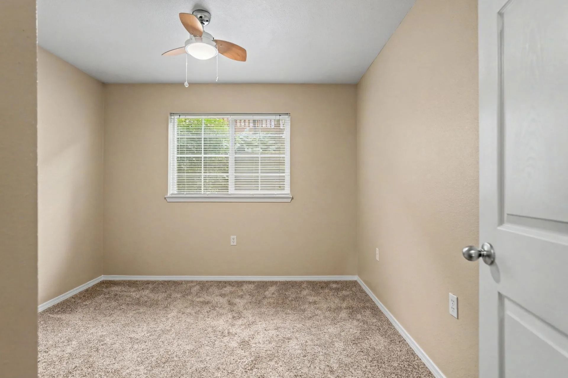 Empty beige bedroom with carpet, ceiling fan, and window blinds.