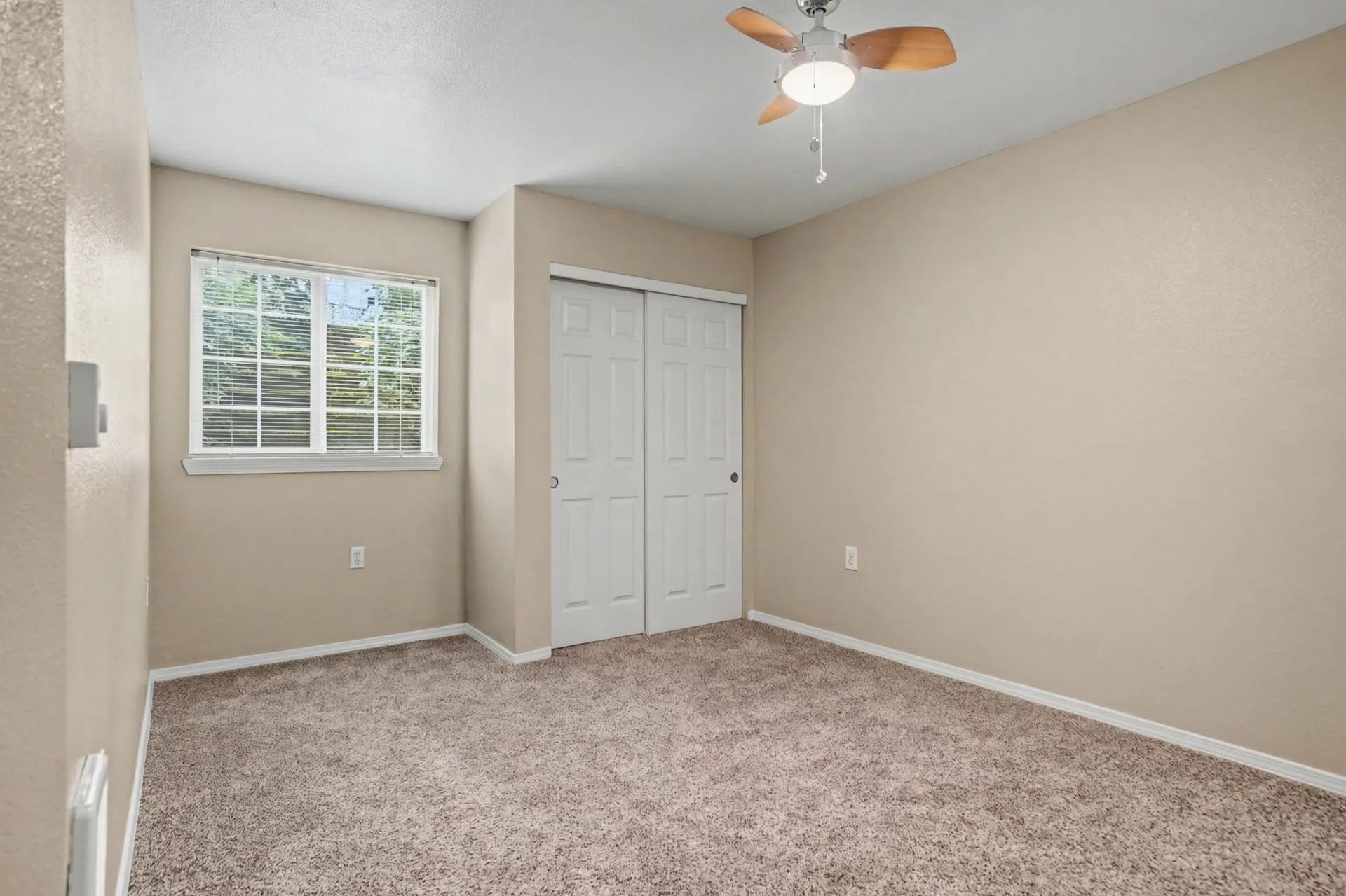 Empty beige bedroom with carpet, window blinds, white closet doors, and ceiling fan.