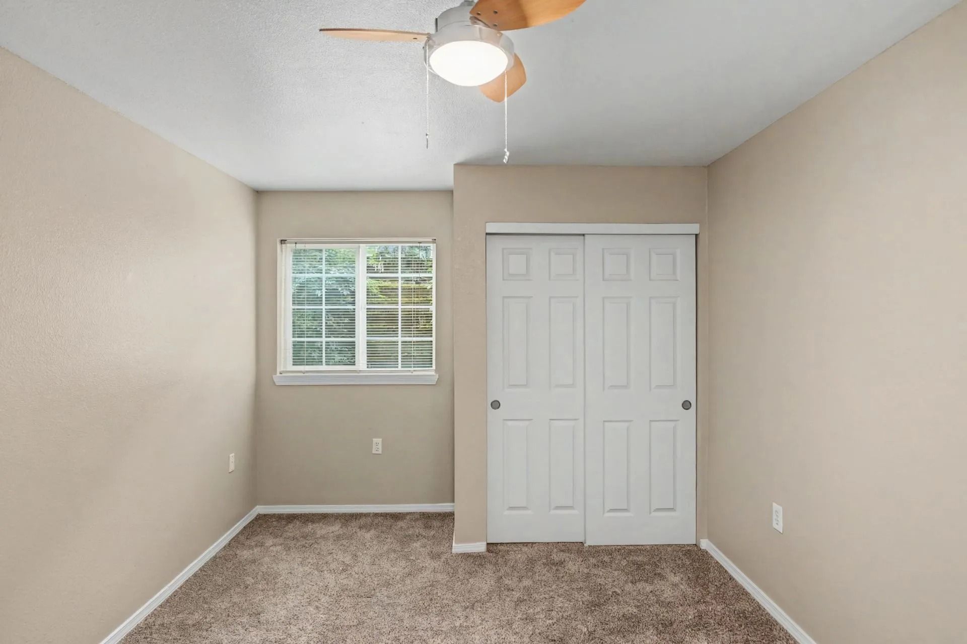 Empty beige bedroom with a window, closet doors, carpet, and ceiling fan light