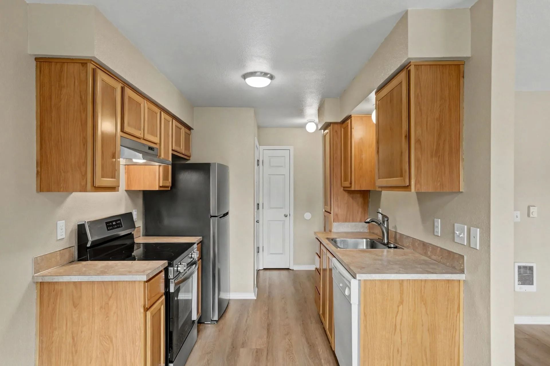 Narrow galley kitchen with wood cabinets, stainless fridge, white walls, and light wood flooring