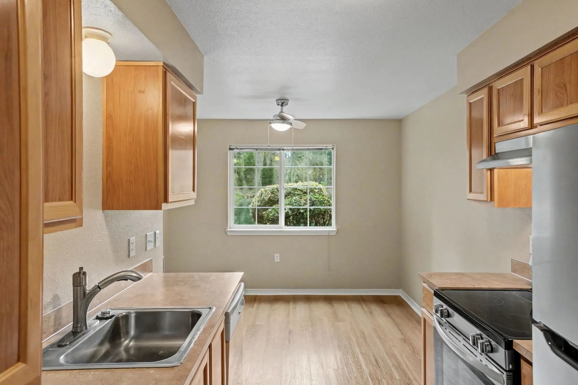 Empty kitchen with wood cabinets, stainless sink and stove, and a window at the far end.