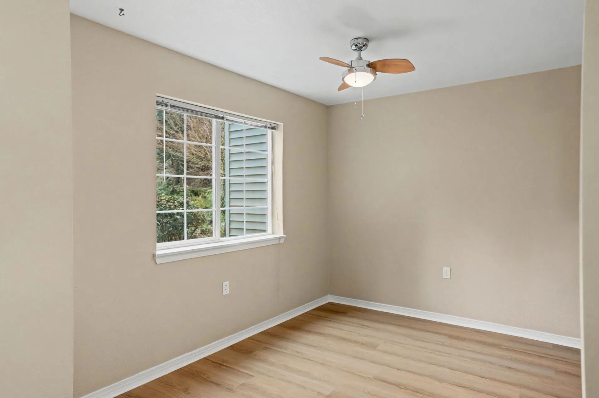 Empty beige bedroom with window, white trim, wood floor, and ceiling fan
