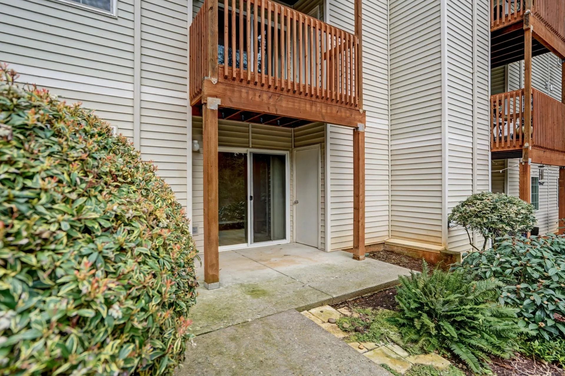 Apartment entrance with wooden balcony above, sliding glass door, and shrubs along the walkway