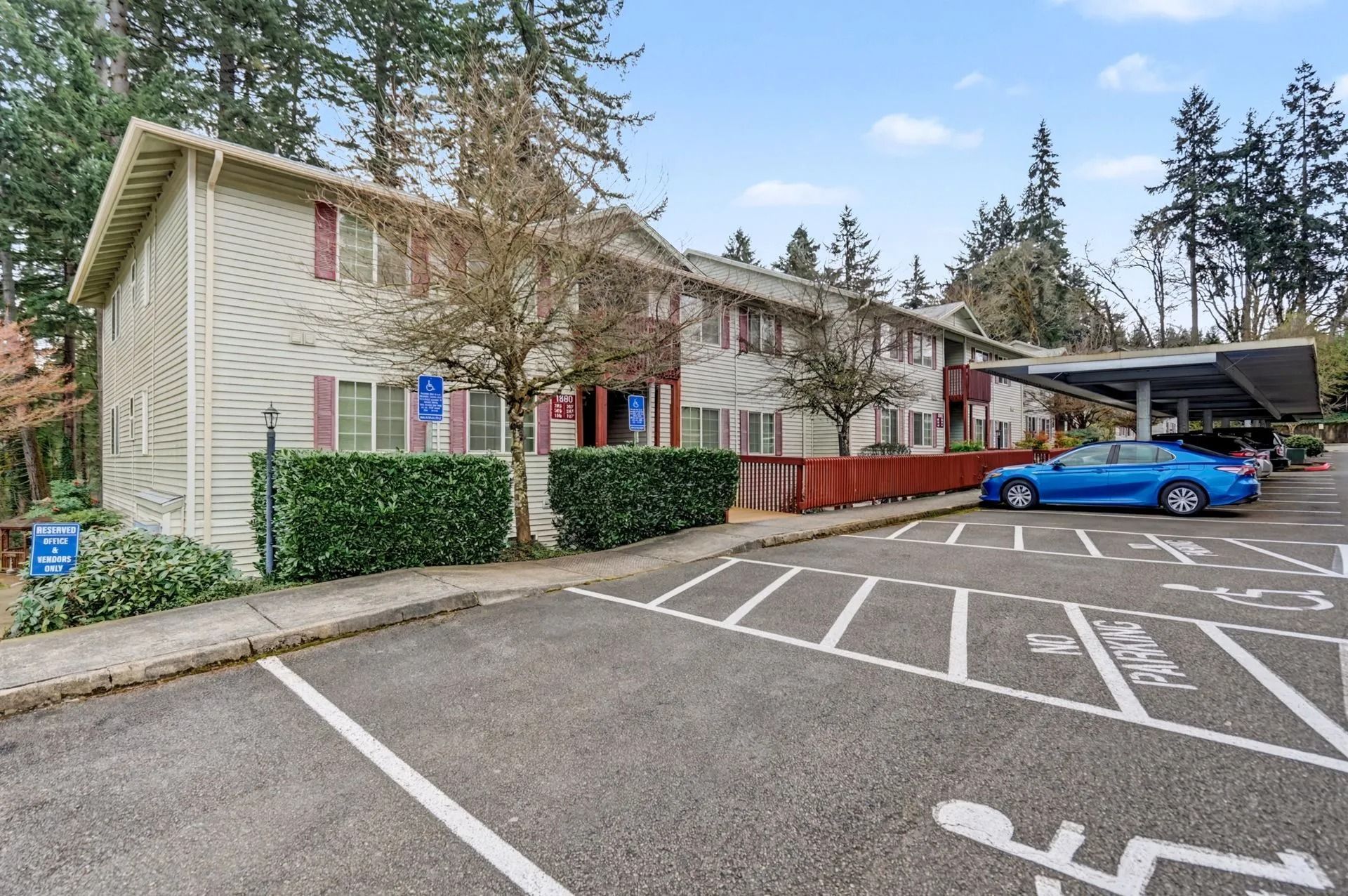 Apartment parking lot with a blue car and red wooden fence beside a beige building and trees