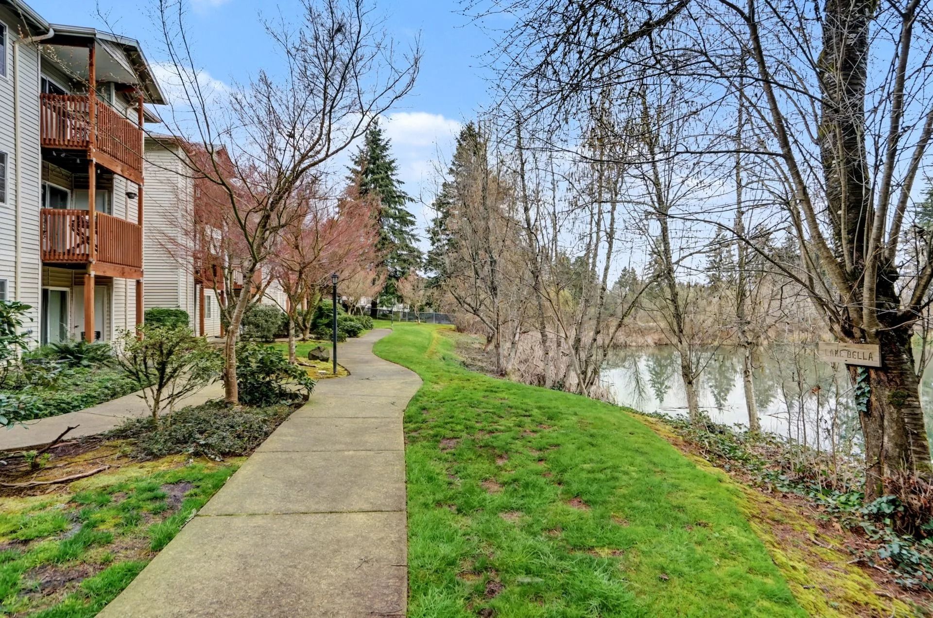 Curved sidewalk beside apartment buildings, green grass, and leafless trees along a calm river or canal