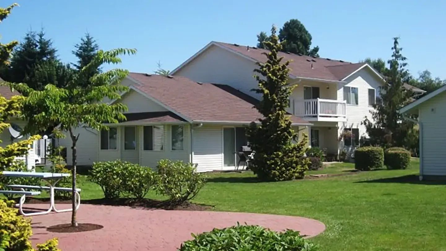 Single-story houses with red roofs beside a green lawn and patio under trees on a sunny day