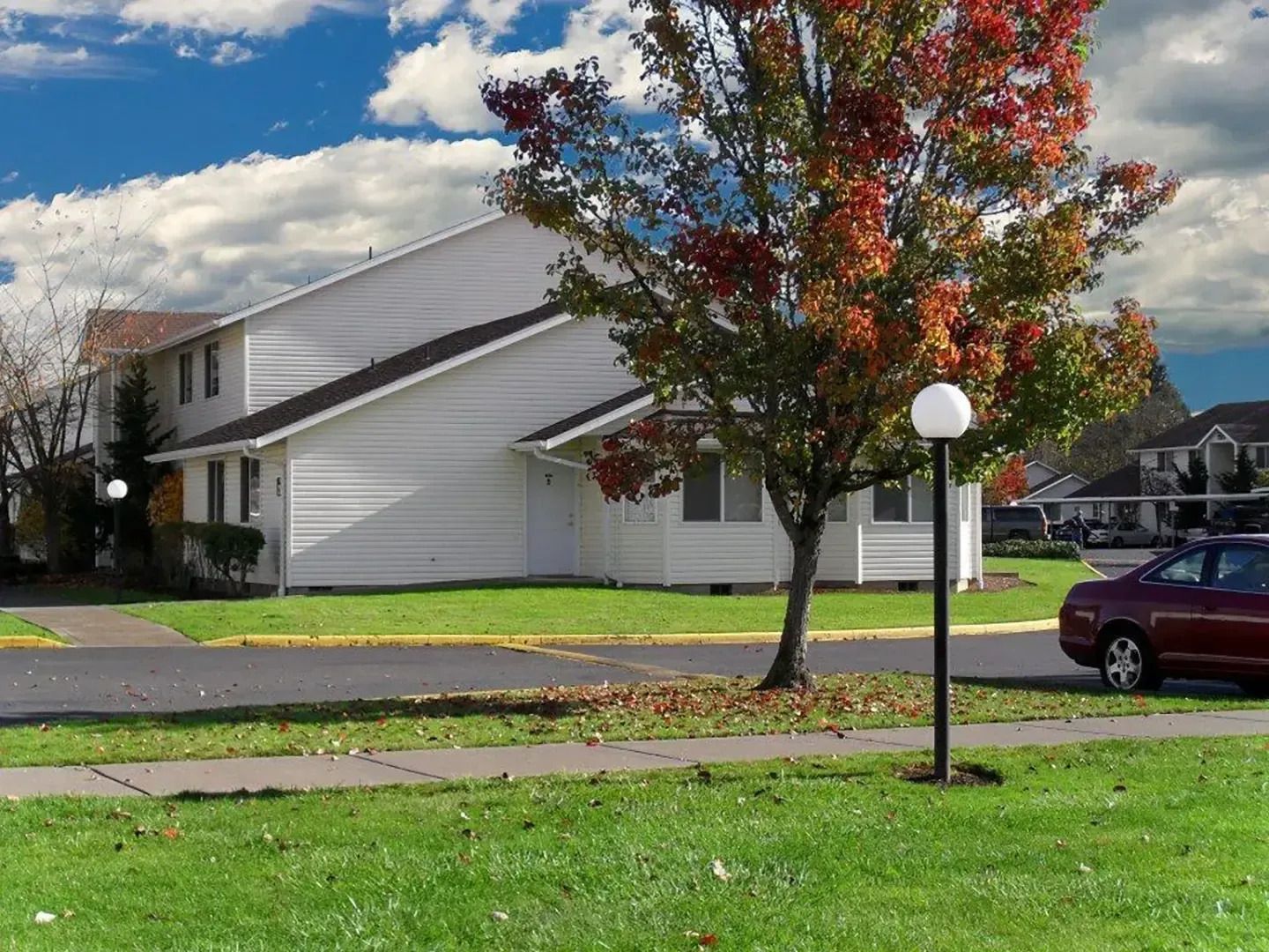 Suburban house with white siding, autumn tree, lawn, and parked maroon car under a cloudy sky
