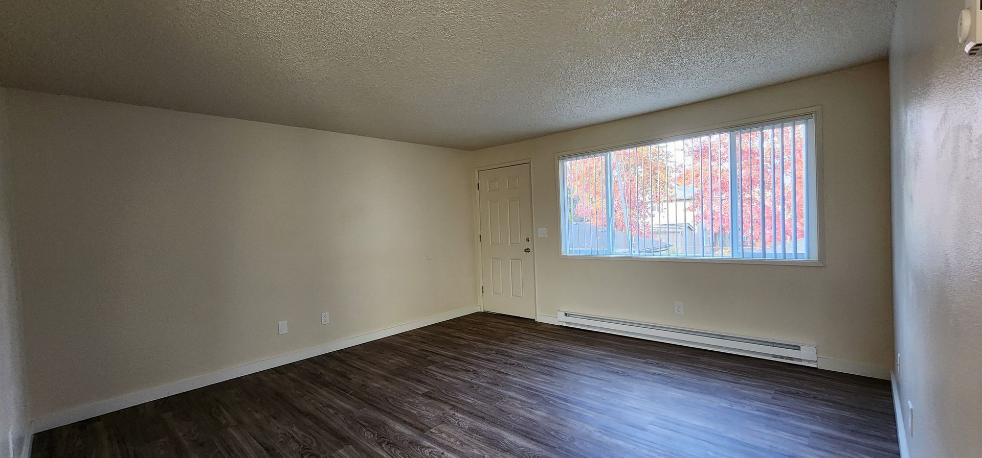 Empty living room with dark wood floors, beige walls, and a window letting in daylight.