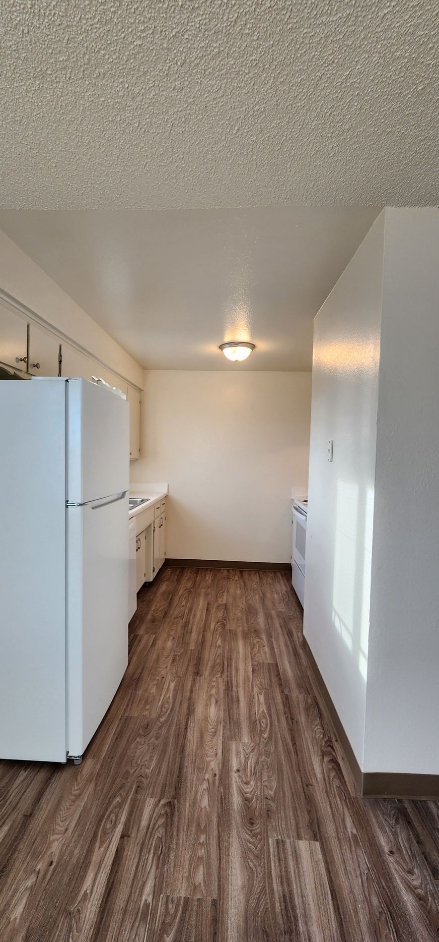 Narrow kitchen with white cabinets and refrigerator, wood-look floor, and a ceiling light.