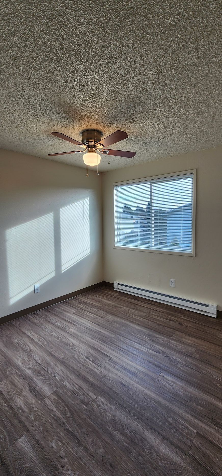 Empty bedroom with dark wood floor, white walls, ceiling fan, and window blinds casting sunlight.