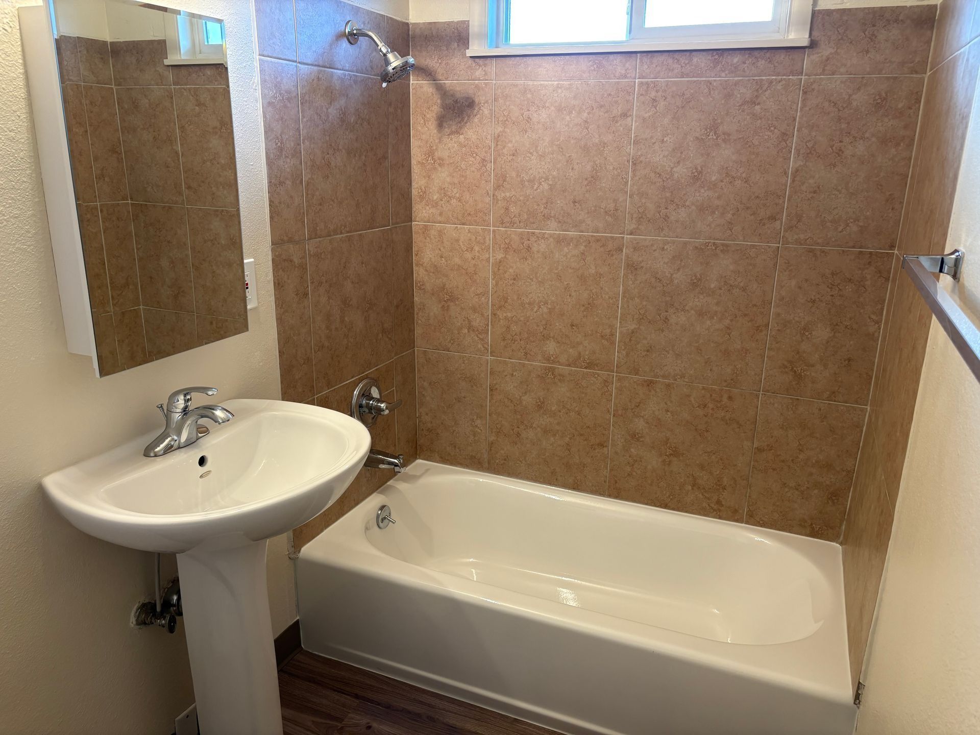 Small bathroom with a white pedestal sink, bathtub, beige tile walls, and a window above the tub.