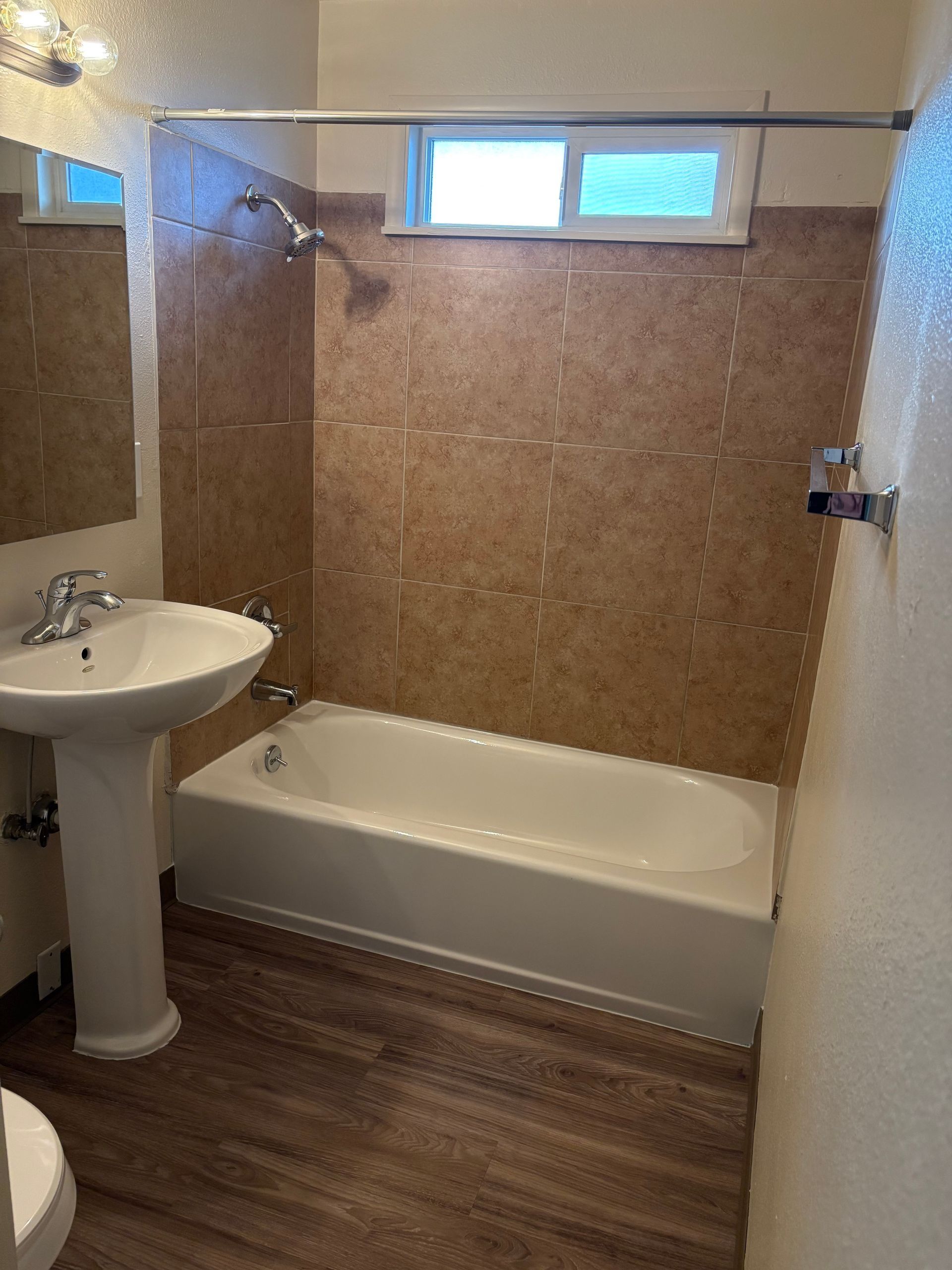 Small bathroom with a pedestal sink, bathtub, window, and brown tile walls.