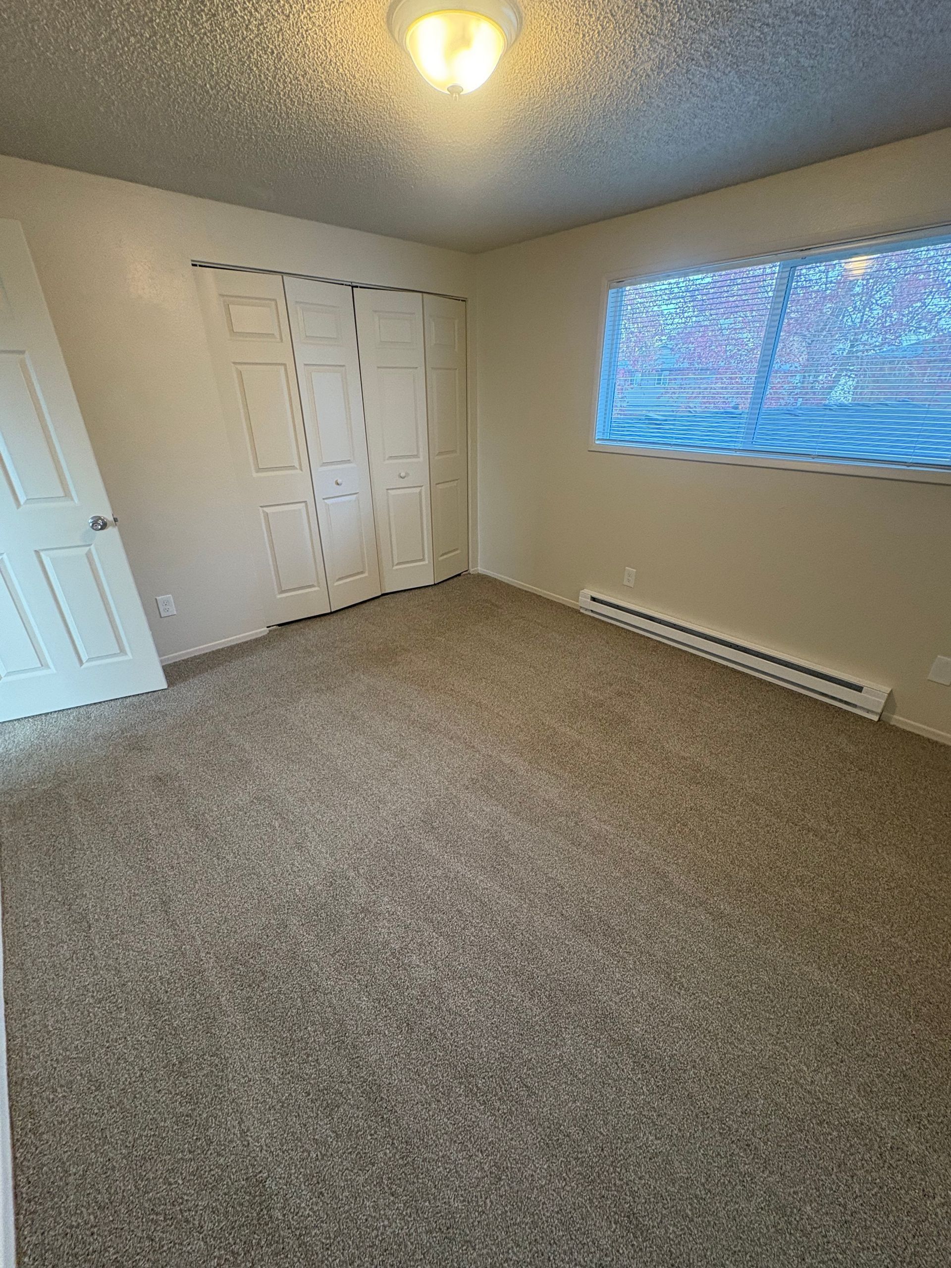 Empty carpeted bedroom with white walls, closet doors, and a window with blinds.