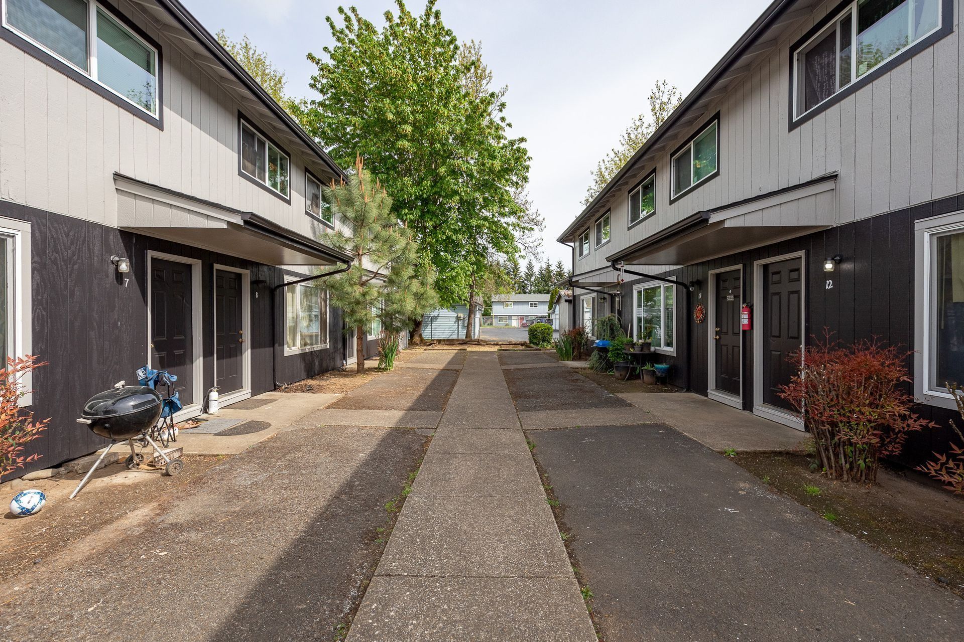 Apartment walkway between two rows of gray townhouses, with a tree and parking area in the distance.