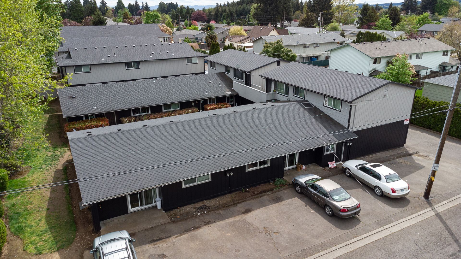 Aerial view of a gray-roofed apartment complex with parked cars and surrounding trees.
