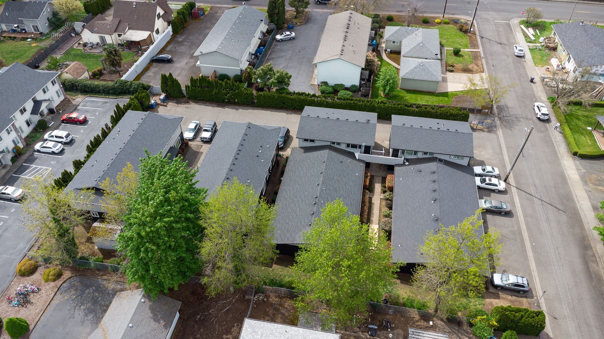 Aerial view of suburban homes with gray roofs, trees, and a street intersection nearby.