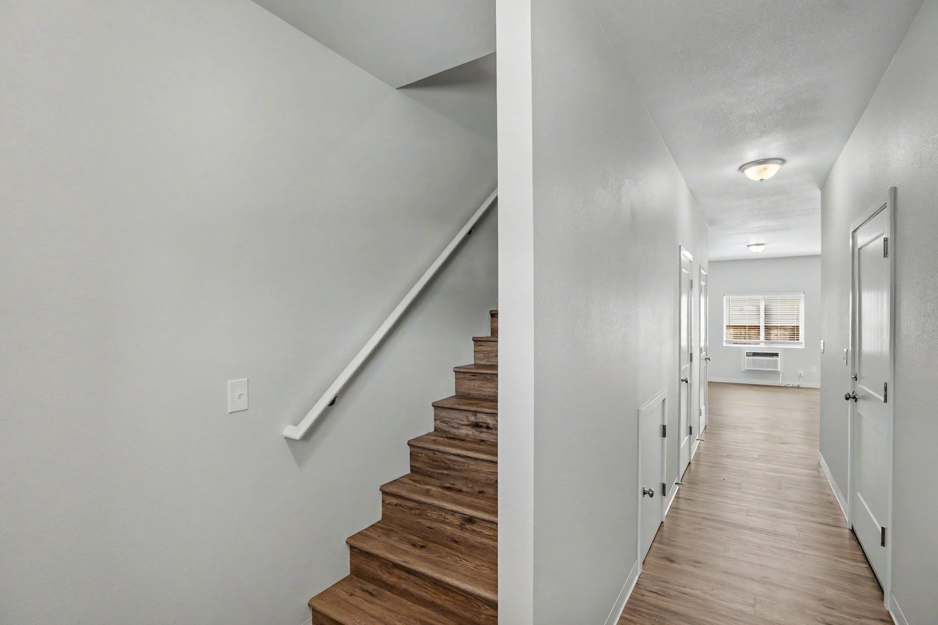Bright hallway with wood floors, white walls, and stairs on the left leading upstairs.