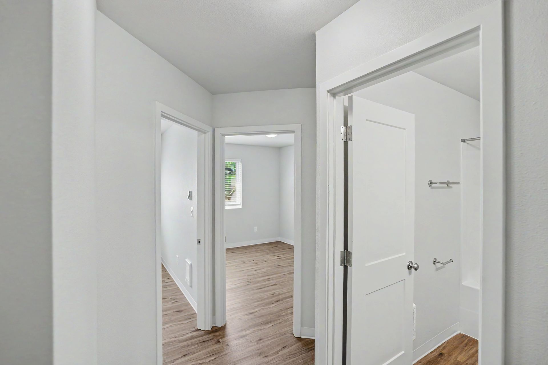 Bright white hallway with open doorways leading to a room with wood floors and a small window.