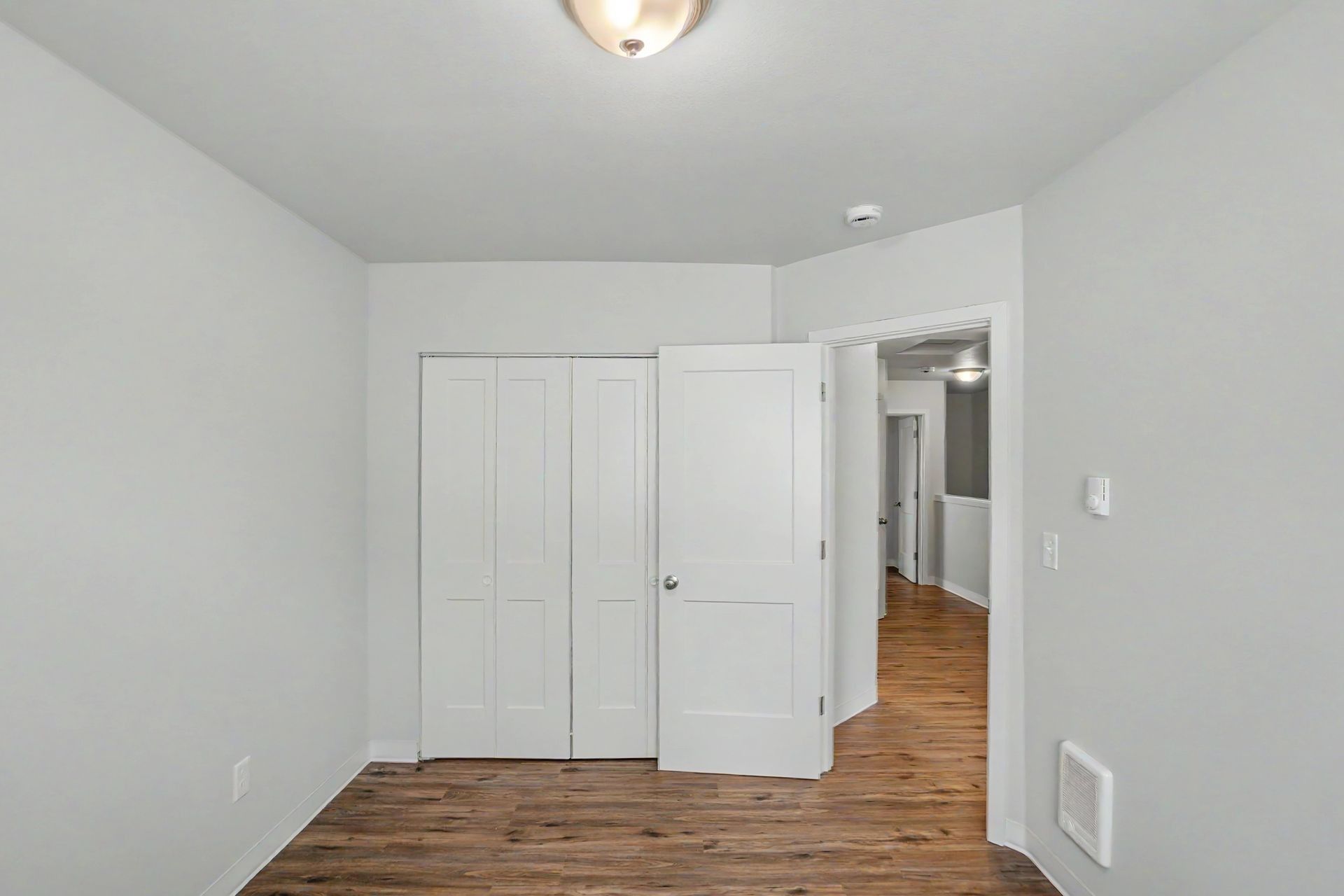 Empty white bedroom with wood flooring and an open door to a hallway