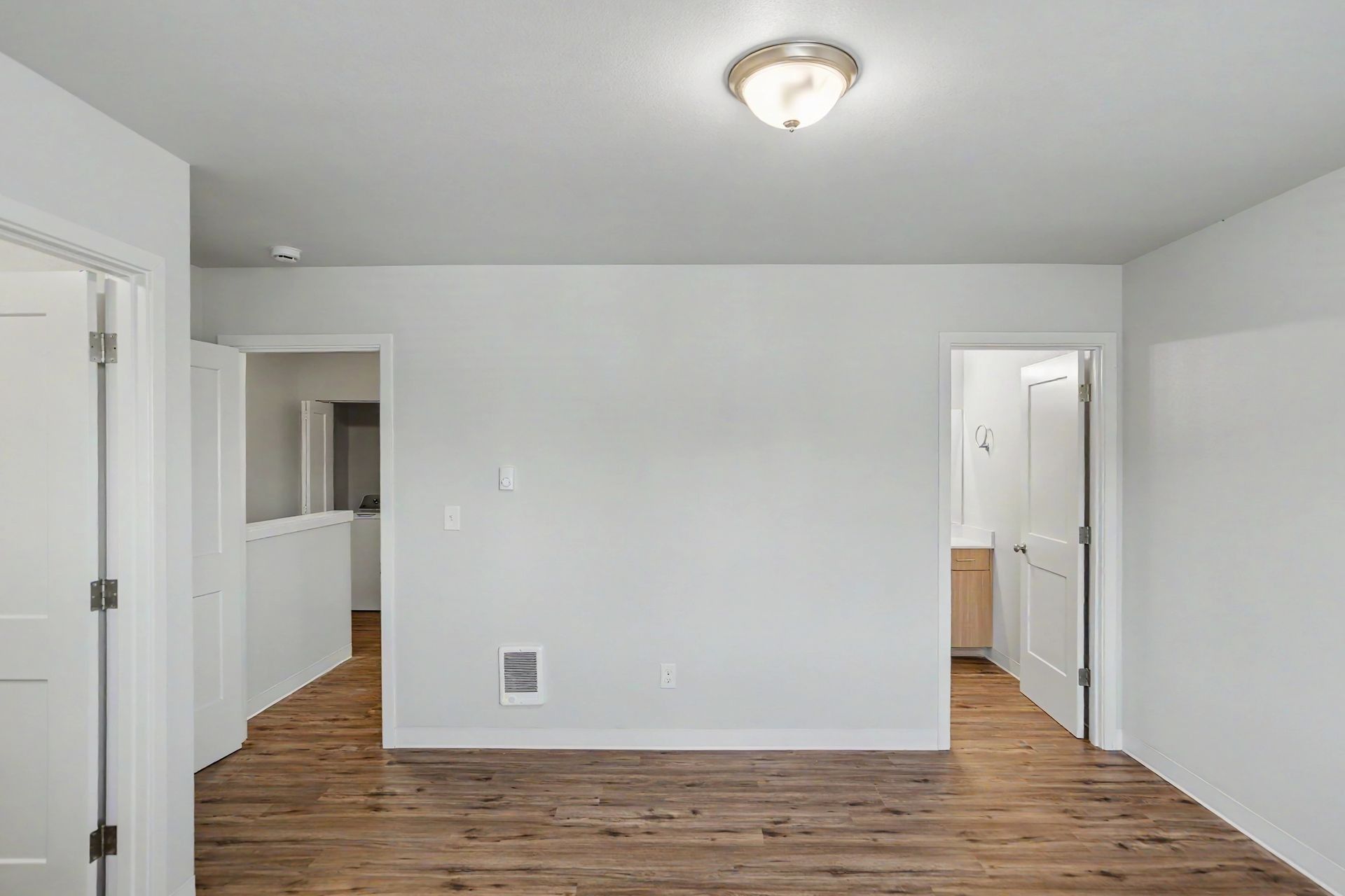 Empty white room with wood floors, ceiling light, and two doorways leading to adjoining spaces