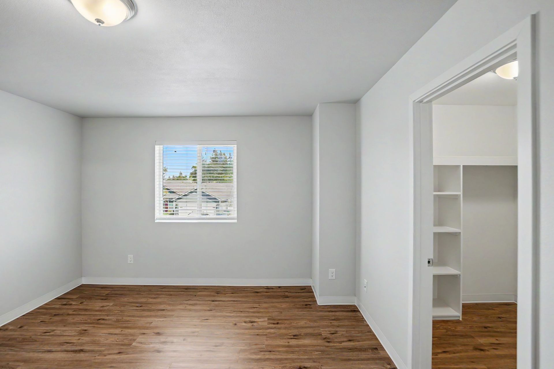 Empty white bedroom with wood floors and a closet opening, lit by a ceiling light and window.