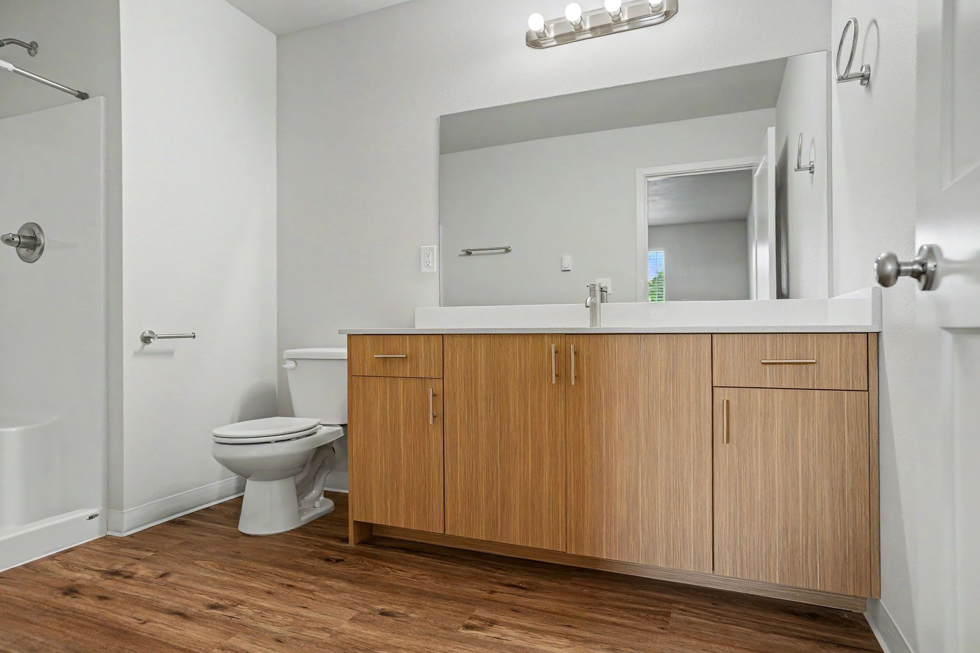 Modern bathroom with wood vanity, large mirror, toilet, and white walls, viewed from the doorway