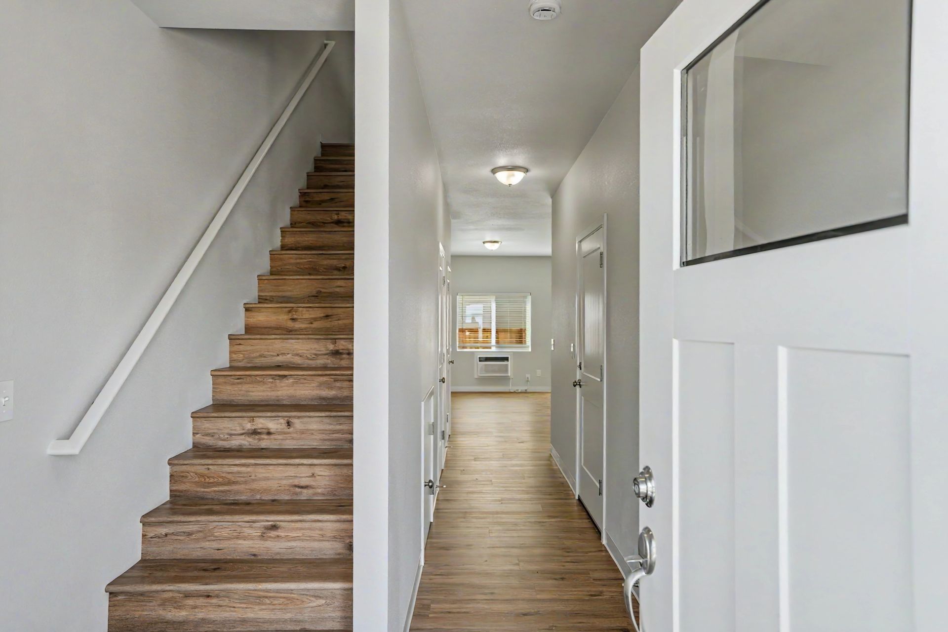 Interior hallway with stairs on the left, white doors, and a wood floor leading to a window.