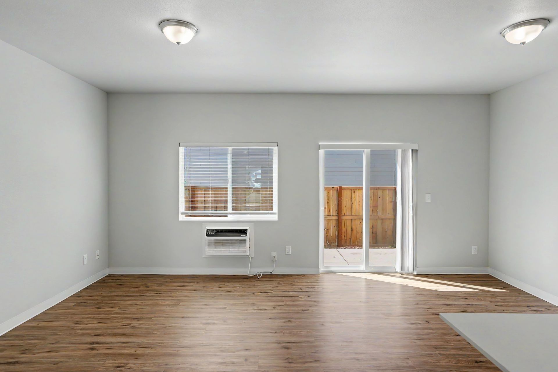 Empty living room with gray walls, hardwood floor, windows, a sliding glass door, and ceiling lights